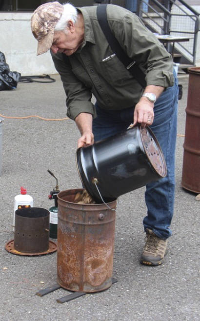 Norm Baker adds chips to one of the smaller versions of burners he has created. The barrels used have holes in the bottom and the lid. The lid has a stovepipe, when burning correctly, it creates no smoke.