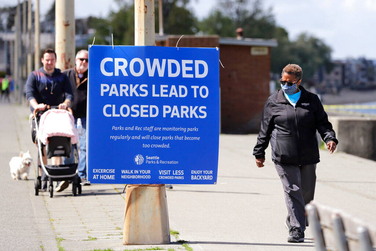 Velma Mullen, right, wears a protective mask as she walks past a sign advising park users to keep physical space between them Monday, April 27, 2020, in Seattle. City guidelines for Seattle parks ask that people stay at least six feet apart, not to congregate and to keep moving to help prevent spread of the coronavirus. (Elaine Thompson/Associated Press)