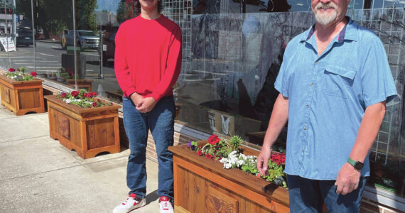 Photo Christi Baron
As part of a recent city beautification project, the aging flower boxes are being replaced, complementing the newly added hanging baskets. Several local businesses have already received the updated boxes, which were crafted at Olympic Corrections Center. Mayor Fletcher had envisioned incorporating tribal art into the project and was introduced to the artwork of Eli Afterbuffalo, a senior at the Quileute Tribal School. Eli submitted his designs, which were recreated by the OCC woodshop and now appear on the front of each flower box.
Pictured are the newly planted boxes in front of Chinook Pharmacy, with Eli Afterbuffalo on the left and Mayor Fletcher on the right.
