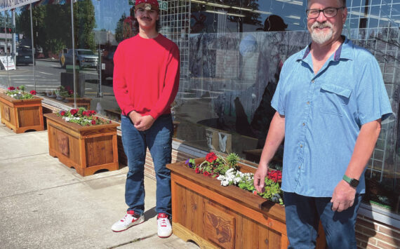 Photo Christi Baron
As part of a recent city beautification project, the aging flower boxes are being replaced, complementing the newly added hanging baskets. Several local businesses have already received the updated boxes, which were crafted at Olympic Corrections Center. Mayor Fletcher had envisioned incorporating tribal art into the project and was introduced to the artwork of Eli Afterbuffalo, a senior at the Quileute Tribal School. Eli submitted his designs, which were recreated by the OCC woodshop and now appear on the front of each flower box.
Pictured are the newly planted boxes in front of Chinook Pharmacy, with Eli Afterbuffalo on the left and Mayor Fletcher on the right.