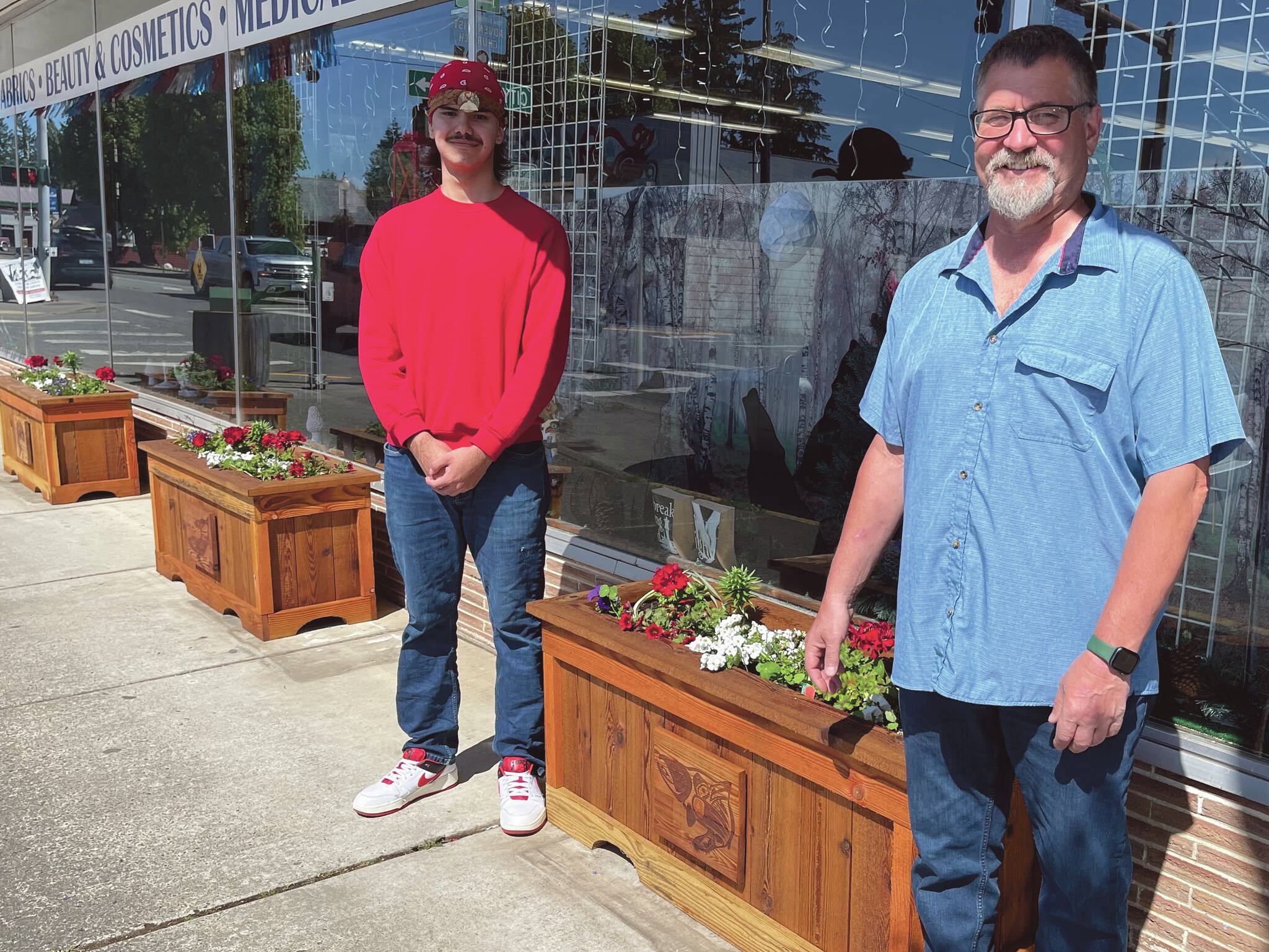 Photo Christi Baron
As part of a recent city beautification project, the aging flower boxes are being replaced, complementing the newly added hanging baskets. Several local businesses have already received the updated boxes, which were crafted at Olympic Corrections Center. Mayor Fletcher had envisioned incorporating tribal art into the project and was introduced to the artwork of Eli Afterbuffalo, a senior at the Quileute Tribal School. Eli submitted his designs, which were recreated by the OCC woodshop and now appear on the front of each flower box.
Pictured are the newly planted boxes in front of Chinook Pharmacy, with Eli Afterbuffalo on the left and Mayor Fletcher on the right.